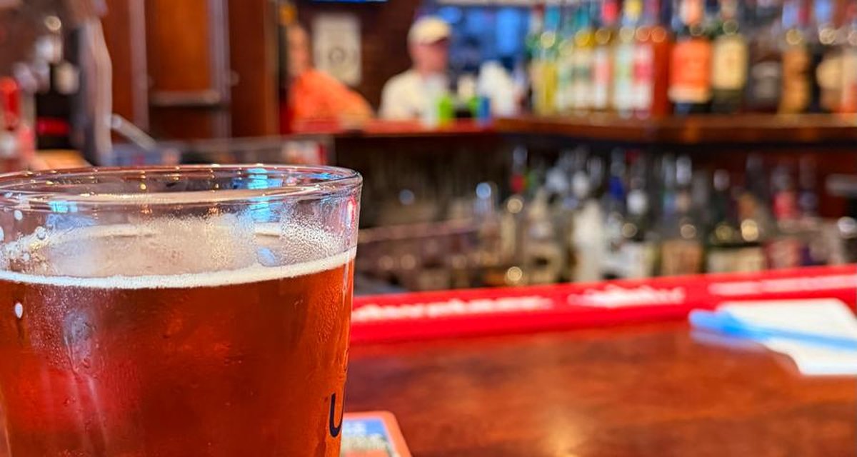 A pint of amber beer on a wooden bar with shelves of bottles behind it