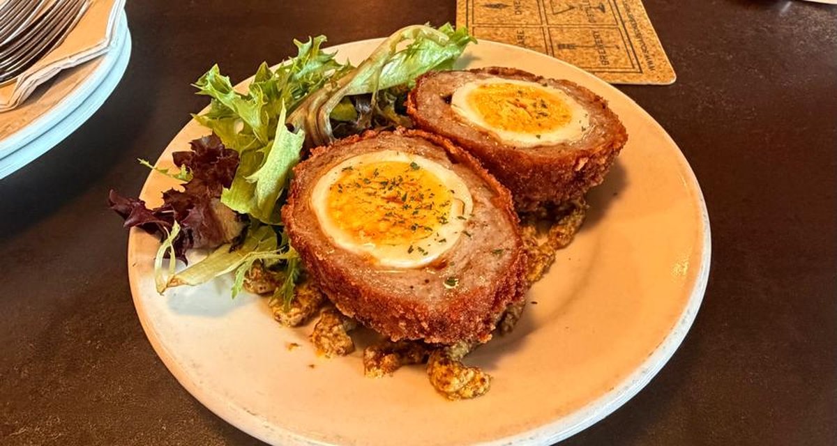 A halved scotch egg on a small plate with greens, on a dark table