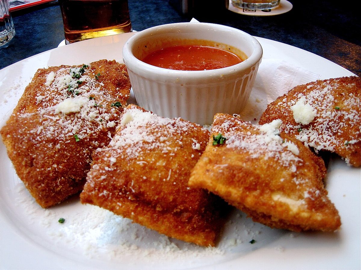 A plate of golden breaded toasted ravioli dusted with grated parmesan, with a small cup of marinara